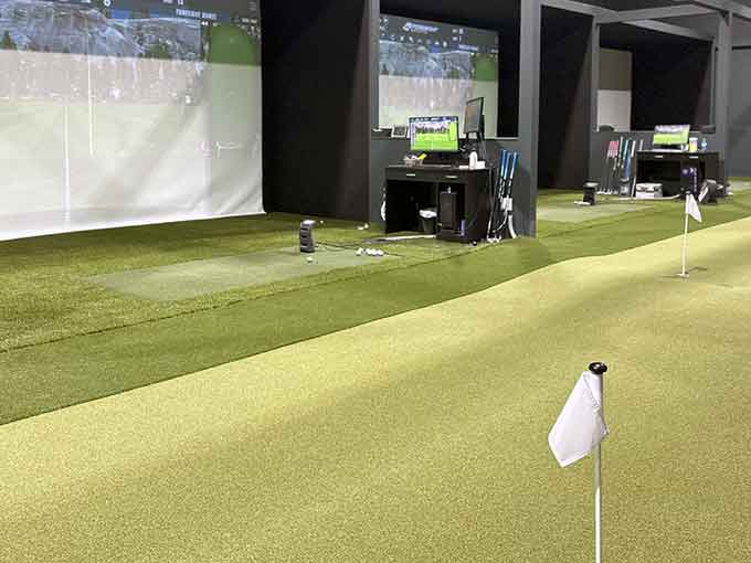 Indoor golf area with synthetic turf, putting greens, and screens.